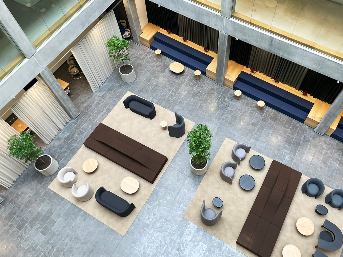 Moderne kontoratrium med siddepladser, der inkluderer sofaer, lænestole og runde borde på beige tæpper, forstærket af DECOPLANT potteplanter, sat på et gråt stengulv mellem glasvægge og gardiner.