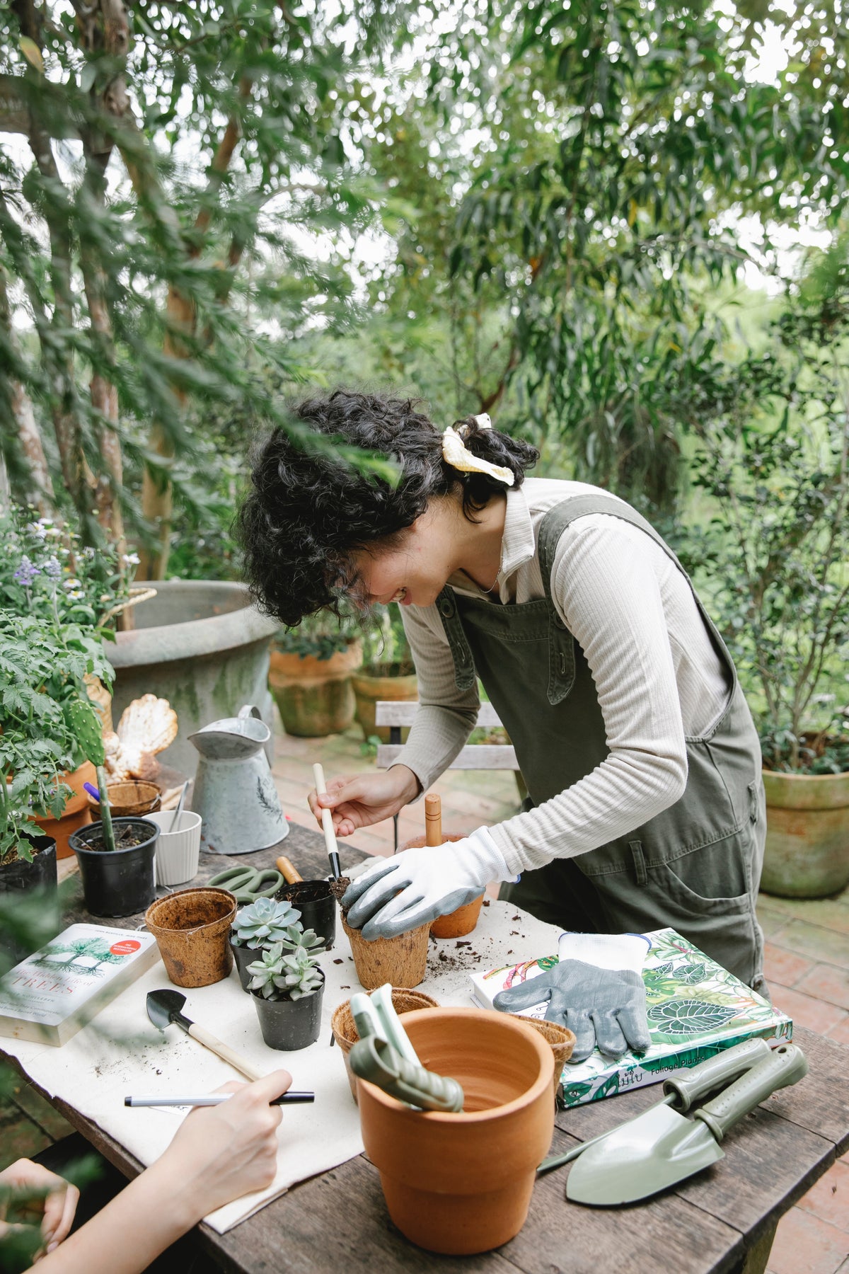 En person i overalls og handsker potter små planter med DECOPLANT forsyninger ved et udendørs bord omgivet af grønt, mens en andens hånd arbejder i nærheden.